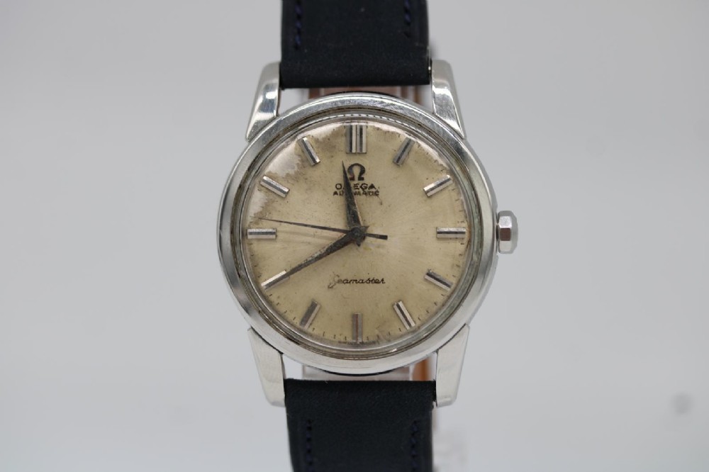 A 1950s stainless steel Omega Seamaster wristwatch featuring a patinated cream dial with silver baton indices and a black leather strap.