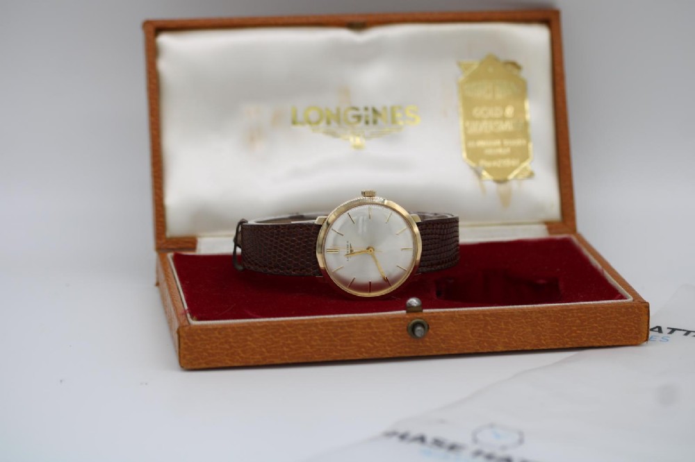 A vintage circular 9ct gold wristwatch by Longines featuring a silver dial with gold baton hour markers and a brown leather strap. View II