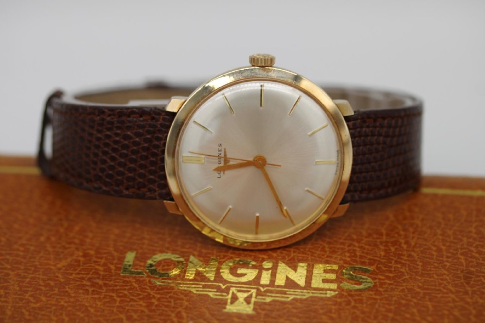 A vintage circular 9ct gold wristwatch by Longines featuring a silver dial with gold baton hour markers and a brown leather strap. View III