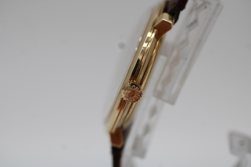 A vintage circular 9ct gold wristwatch by Longines featuring a silver dial with gold baton hour markers and a brown leather strap. View VI