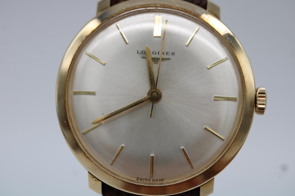 A vintage circular 9ct gold wristwatch by Longines featuring a silver dial with gold baton hour markers and a brown leather strap. View VIII
