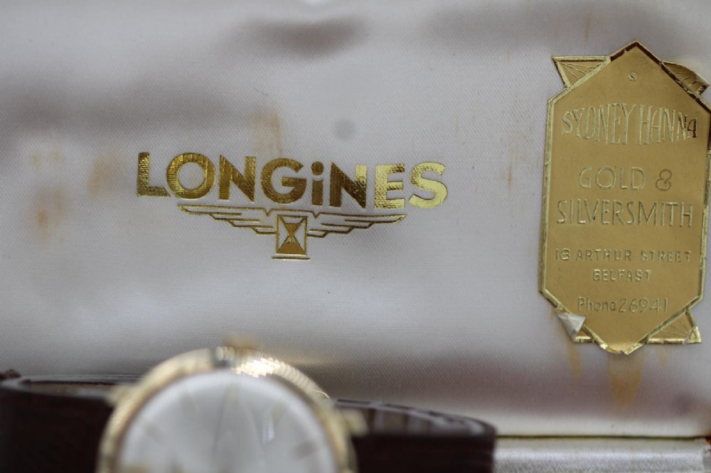 A vintage circular 9ct gold wristwatch by Longines featuring a silver dial with gold baton hour markers and a brown leather strap. View X