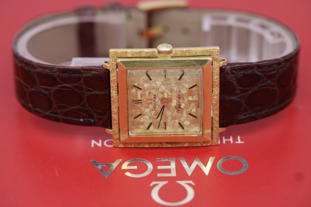 A 1960s Omega Geneve manual wind wristwatch featuring an 18ct yellow gold cushion-shaped case, a dark brown dial with gold Roman numerals at twelve and six, and a brown leather strap.