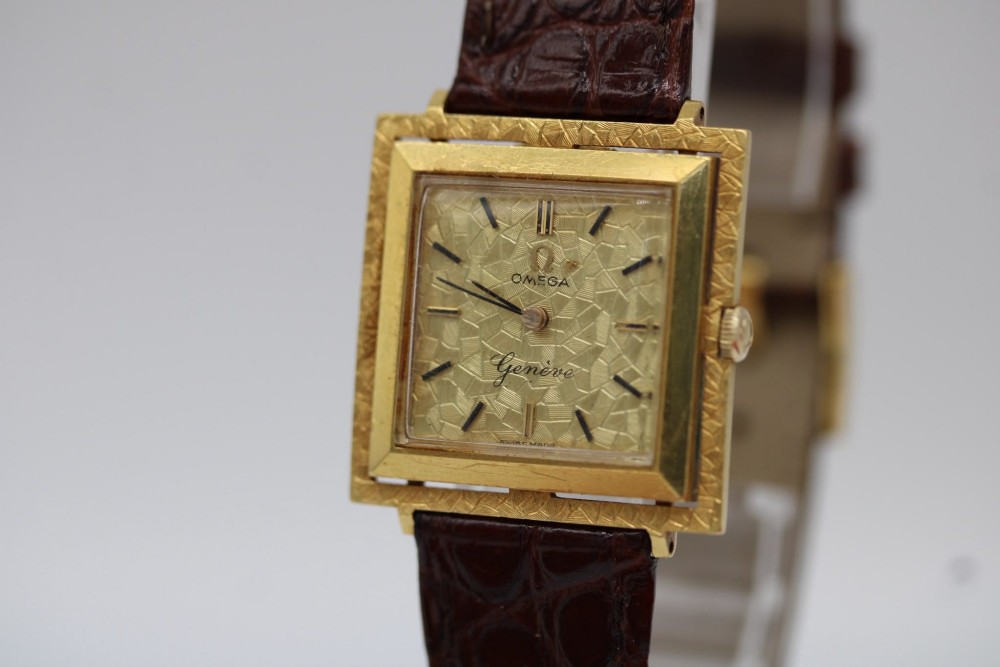 A 1960s Omega Geneve manual wind wristwatch featuring an 18ct yellow gold cushion-shaped case, a dark brown dial with gold Roman numerals at twelve and six, and a brown leather strap. View V