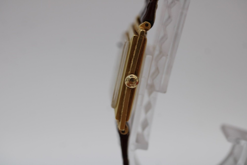 A 1960s Omega Geneve manual wind wristwatch featuring an 18ct yellow gold cushion-shaped case, a dark brown dial with gold Roman numerals at twelve and six, and a brown leather strap. View VI
