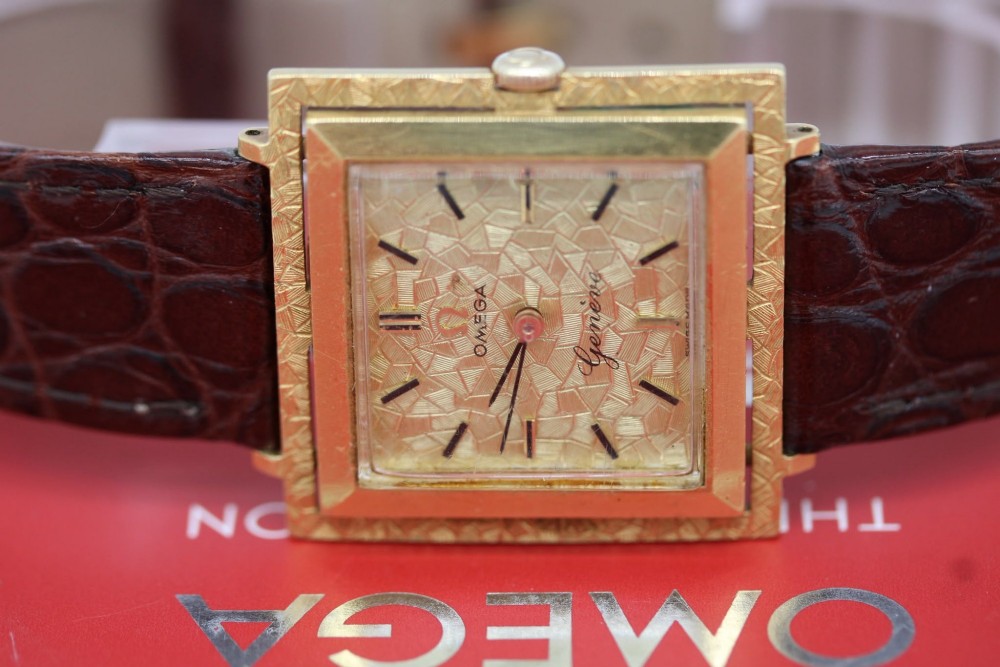 A 1960s Omega Geneve manual wind wristwatch featuring an 18ct yellow gold cushion-shaped case, a dark brown dial with gold Roman numerals at twelve and six, and a brown leather strap. View VII