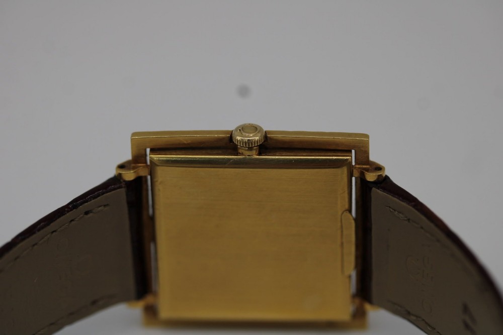 A 1960s Omega Geneve manual wind wristwatch featuring an 18ct yellow gold cushion-shaped case, a dark brown dial with gold Roman numerals at twelve and six, and a brown leather strap. View VIII