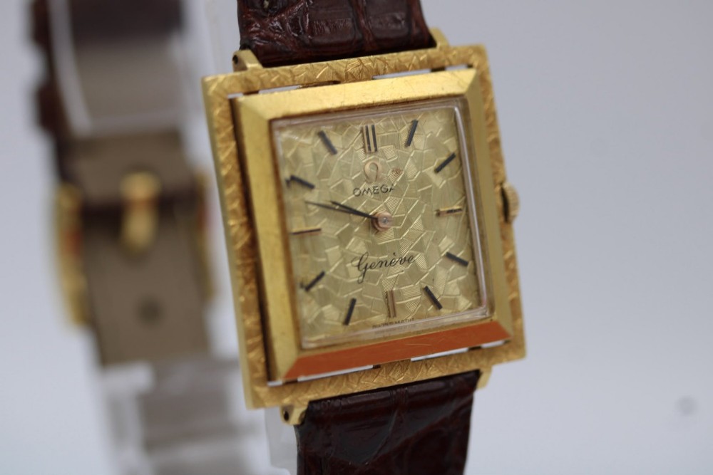 A 1960s Omega Geneve manual wind wristwatch featuring an 18ct yellow gold cushion-shaped case, a dark brown dial with gold Roman numerals at twelve and six, and a brown leather strap. View X