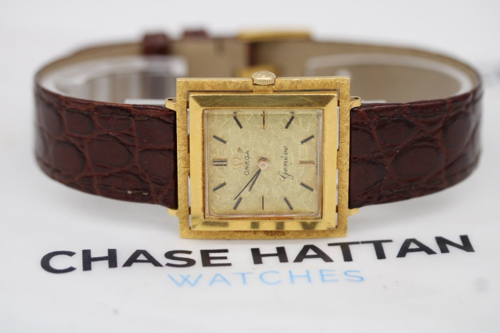 A 1960s Omega Geneve manual wind wristwatch featuring an 18ct yellow gold cushion-shaped case, a dark brown dial with gold Roman numerals at twelve and six, and a brown leather strap. View XI