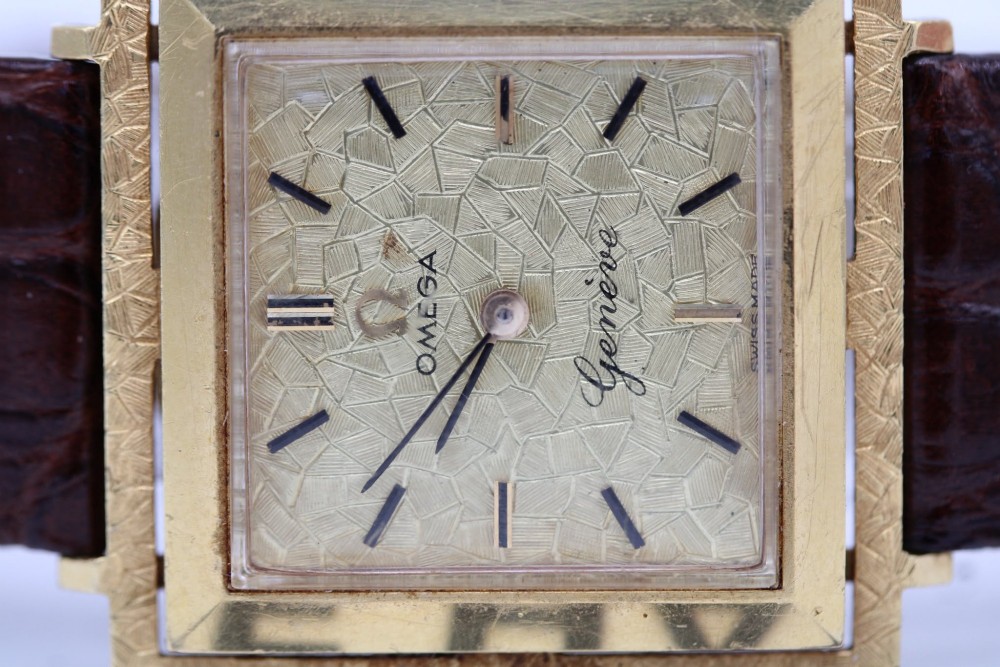 A 1960s Omega Geneve manual wind wristwatch featuring an 18ct yellow gold cushion-shaped case, a dark brown dial with gold Roman numerals at twelve and six, and a brown leather strap. View XII