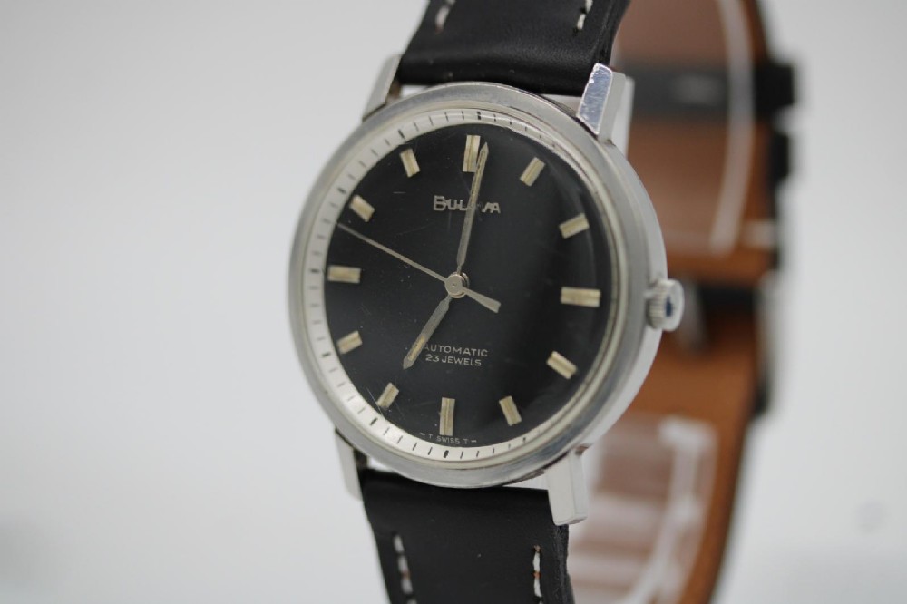 A vintage Bulova automatic wristwatch featuring a black dial with silver baton hour markers and a black leather strap with white stitching. View IV
