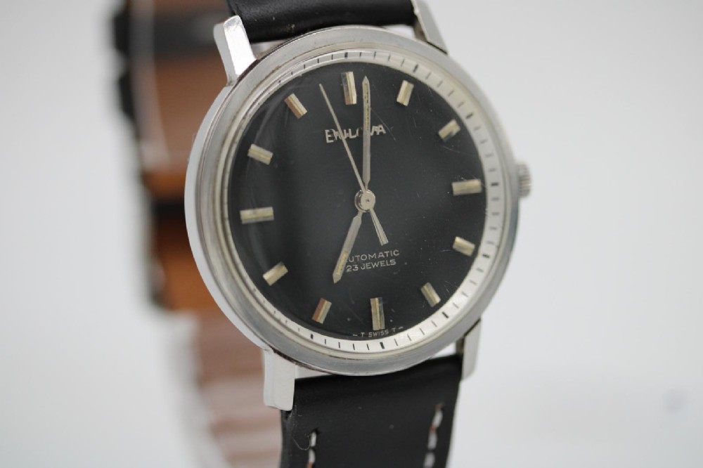 A vintage Bulova automatic wristwatch featuring a black dial with silver baton hour markers and a black leather strap with white stitching. View V