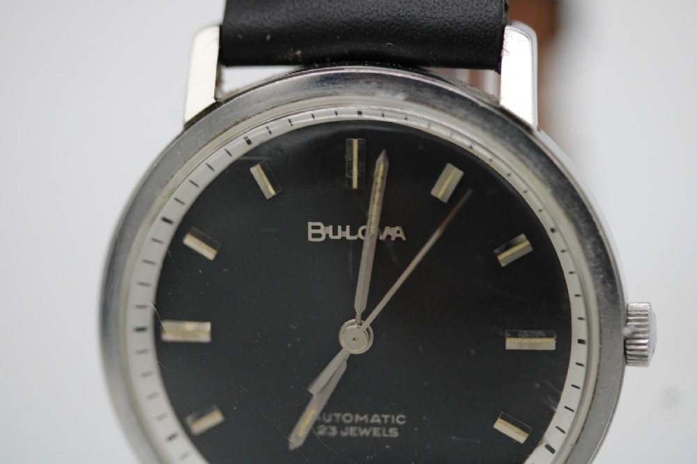 A vintage Bulova automatic wristwatch featuring a black dial with silver baton hour markers and a black leather strap with white stitching. View VI