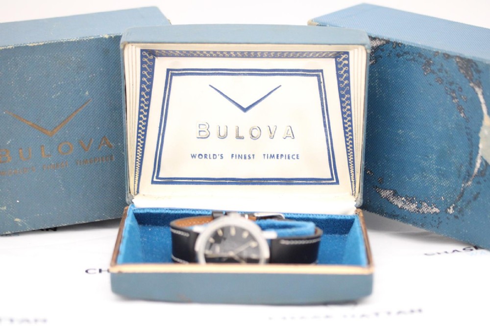 A vintage Bulova automatic wristwatch featuring a black dial with silver baton hour markers and a black leather strap with white stitching. View X