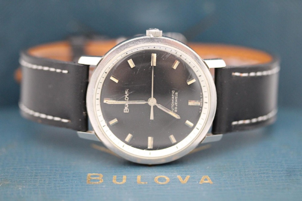 A vintage Bulova automatic wristwatch featuring a black dial with silver baton hour markers and a black leather strap with white stitching. View XI