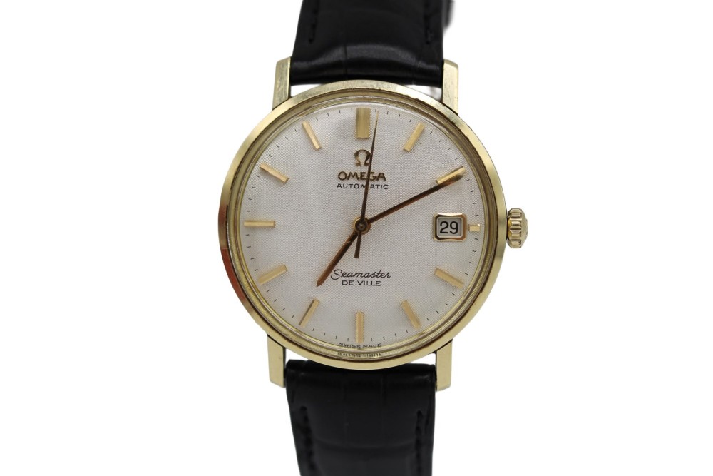 A vintage gold-plated Omega Seamaster DeVille automatic wristwatch with a white textured dial, gold baton indices, date window, and black leather strap.