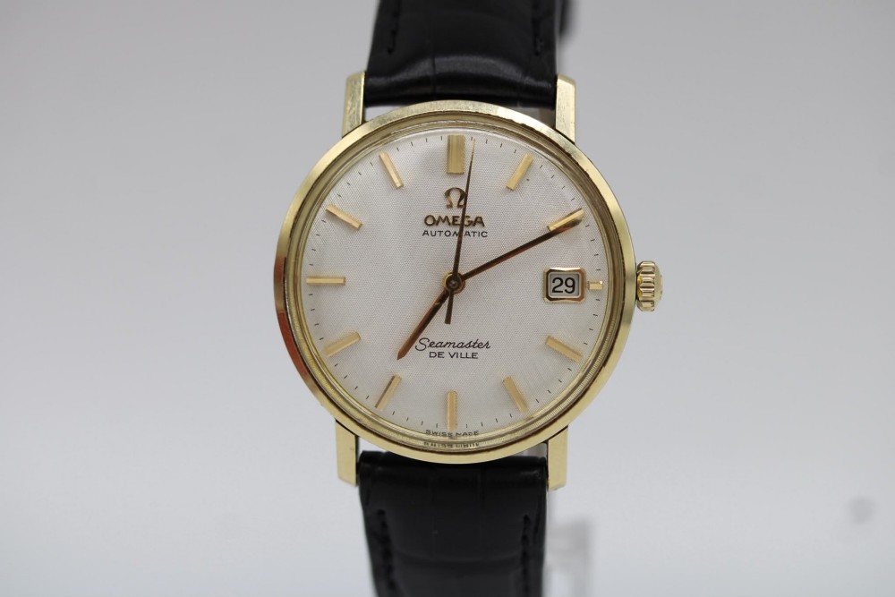 A vintage gold-plated Omega Seamaster DeVille automatic wristwatch with a white textured dial, gold baton indices, date window, and black leather strap. View IV