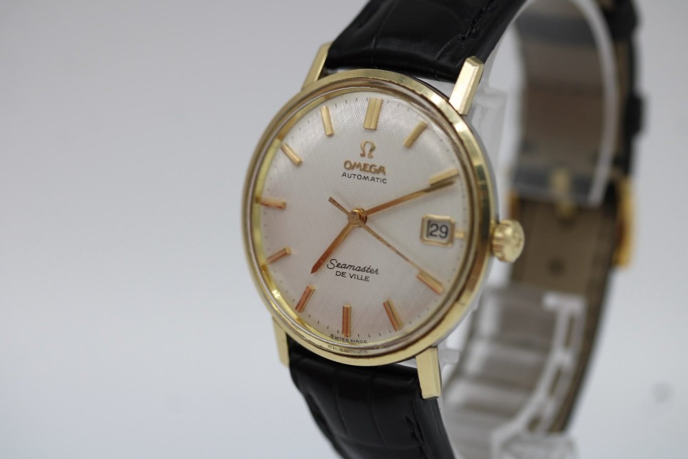 A vintage gold-plated Omega Seamaster DeVille automatic wristwatch with a white textured dial, gold baton indices, date window, and black leather strap. View V