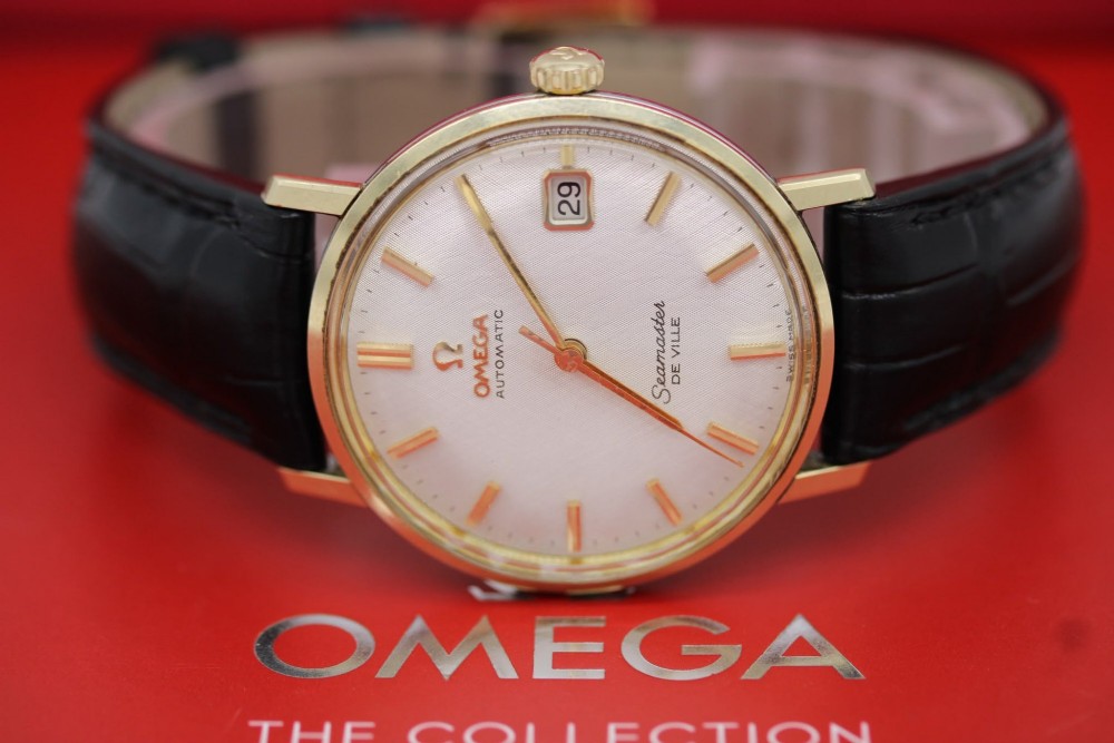 A vintage gold-plated Omega Seamaster DeVille automatic wristwatch with a white textured dial, gold baton indices, date window, and black leather strap. View VI