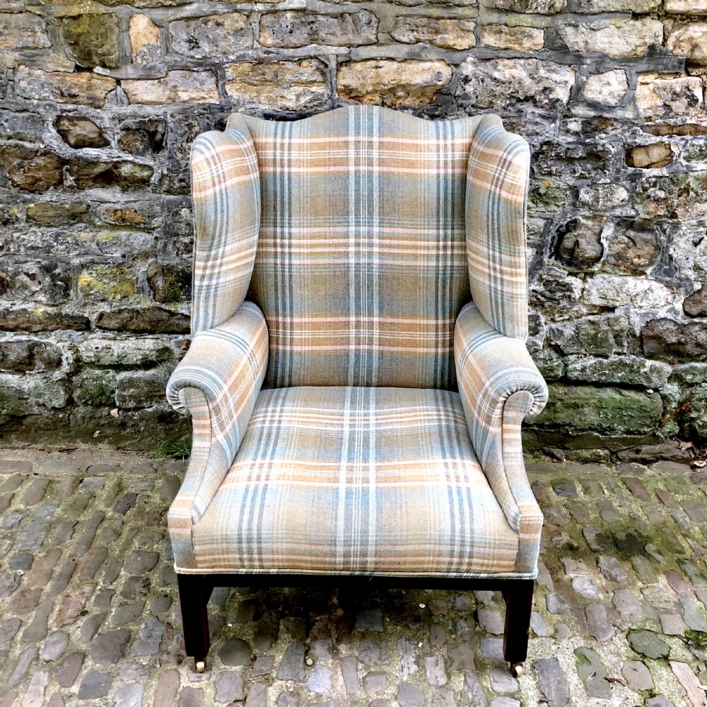 A George III mahogany wing armchair upholstered in a blue, ochre, and cream tartan wool fabric with out-scrolled arms and square legs on brass castors.