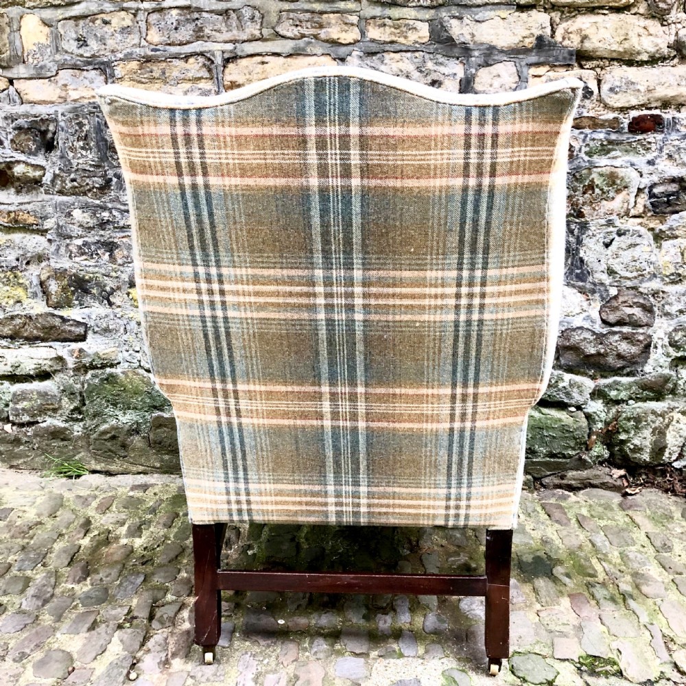 A George III mahogany wing armchair upholstered in a blue, ochre, and cream tartan wool fabric with out-scrolled arms and square legs on brass castors. View IV