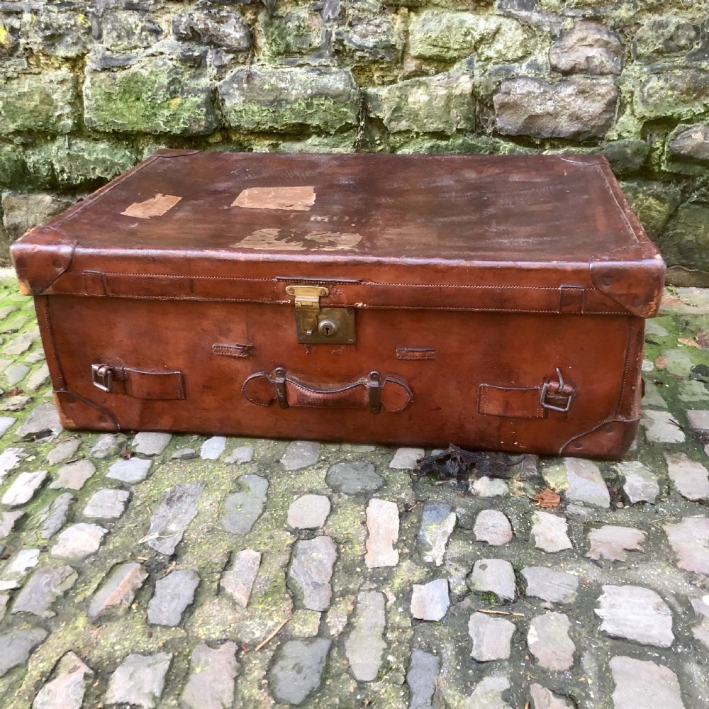 An early 20th-century warm brown leather steamer trunk with stitched corner protectors, leather straps, and a central brass lock mechanism.