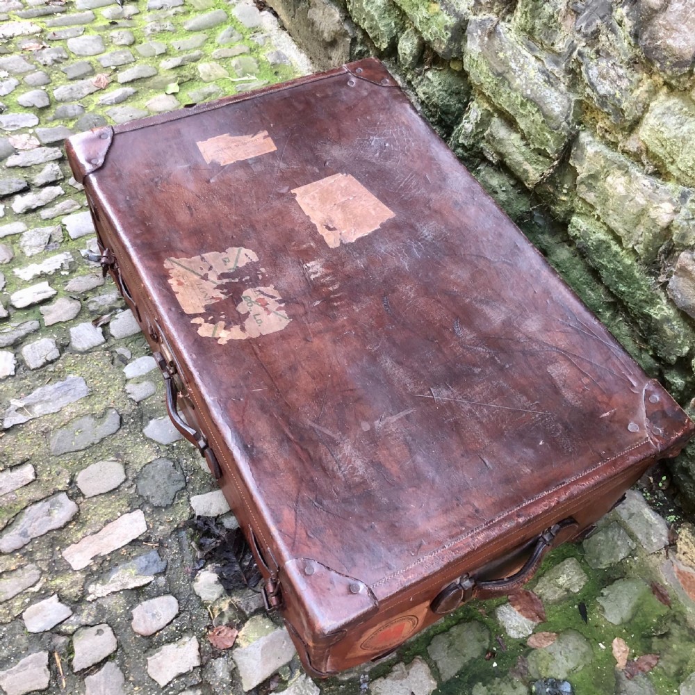 An early 20th-century warm brown leather steamer trunk with stitched corner protectors, leather straps, and a central brass lock mechanism. View V