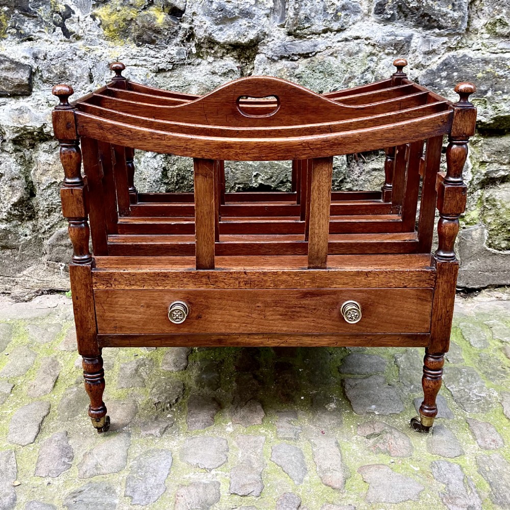 A George III mahogany Canterbury featuring four curved slatted compartments, turned corner posts topped with finials, a single base drawer with brass knobs, and turned legs on brass castors.