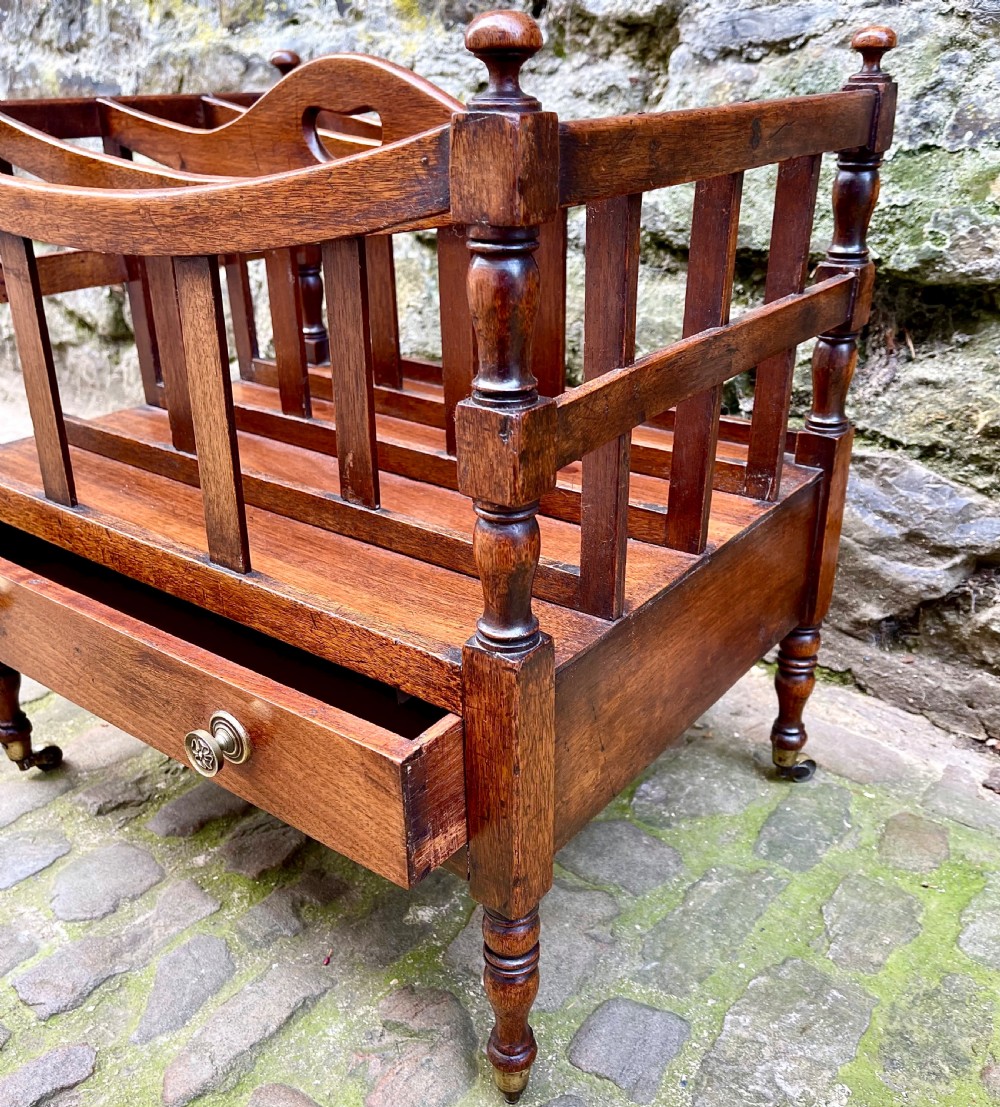 A George III mahogany Canterbury featuring four curved slatted compartments, turned corner posts topped with finials, a single base drawer with brass knobs, and turned legs on brass castors. View III