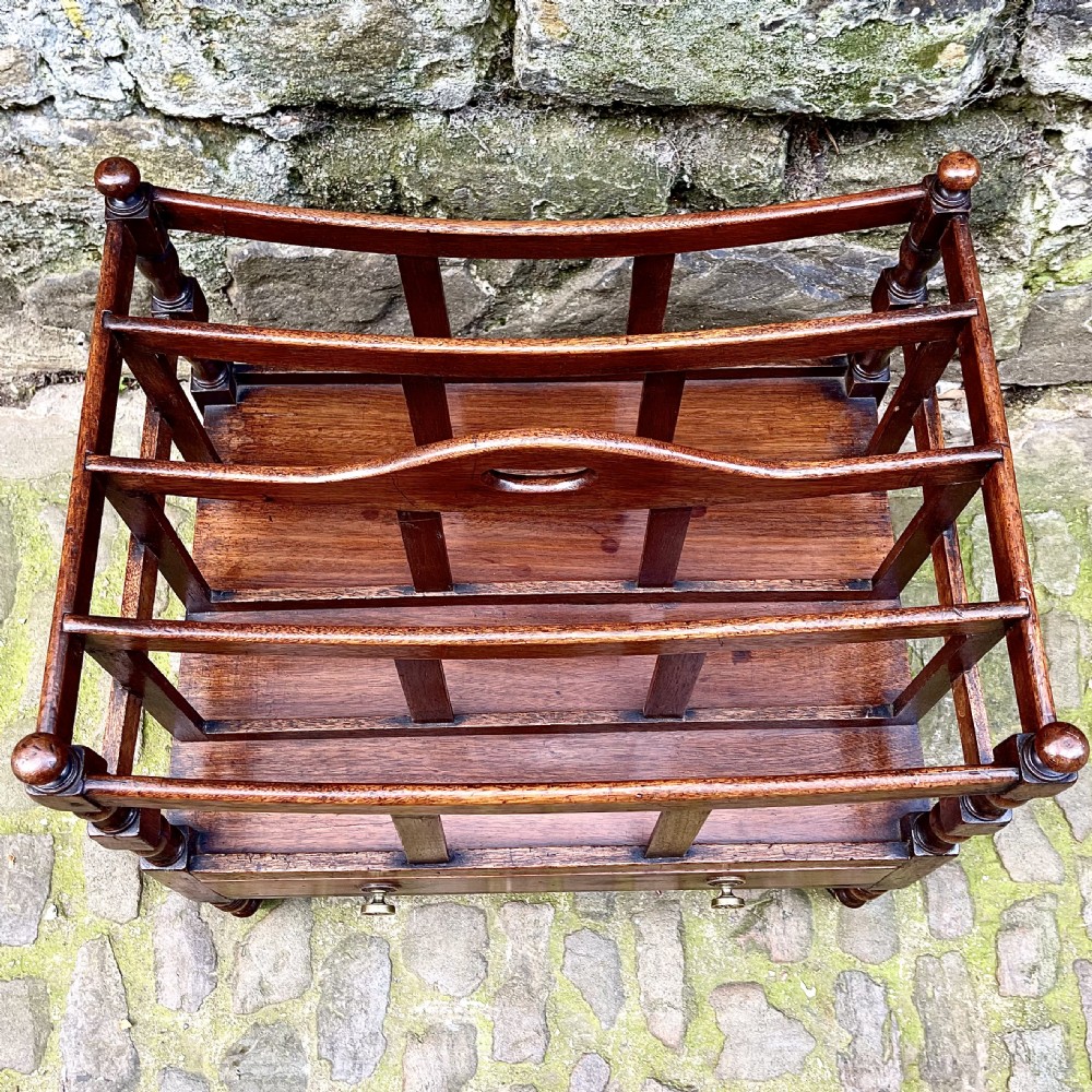 A George III mahogany Canterbury featuring four curved slatted compartments, turned corner posts topped with finials, a single base drawer with brass knobs, and turned legs on brass castors. View IV