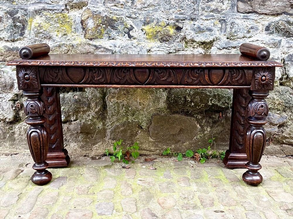 A mid-Victorian dark oak window seat featuring a carved relief frieze, turned front legs, and rolled end supports.