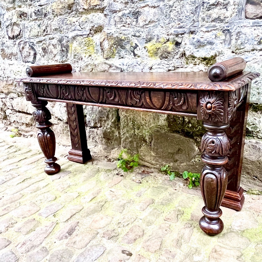 A mid-Victorian dark oak window seat featuring a carved relief frieze, turned front legs, and rolled end supports. View IV