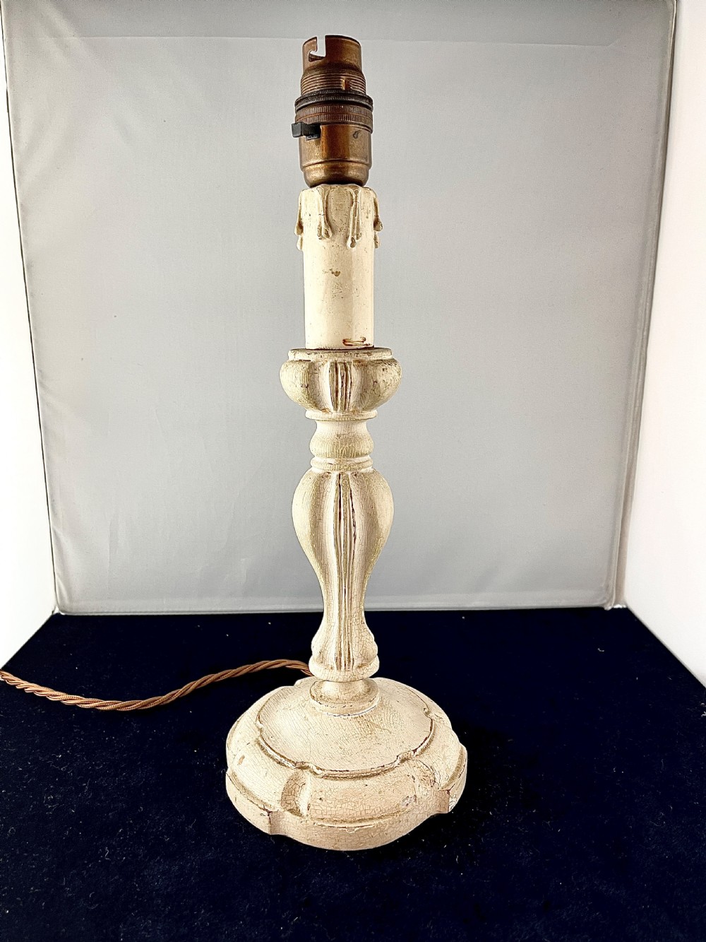 An early 20th century turned wood candlestick table lamp featuring a distressed cream painted finish and a brass bulb fitting.