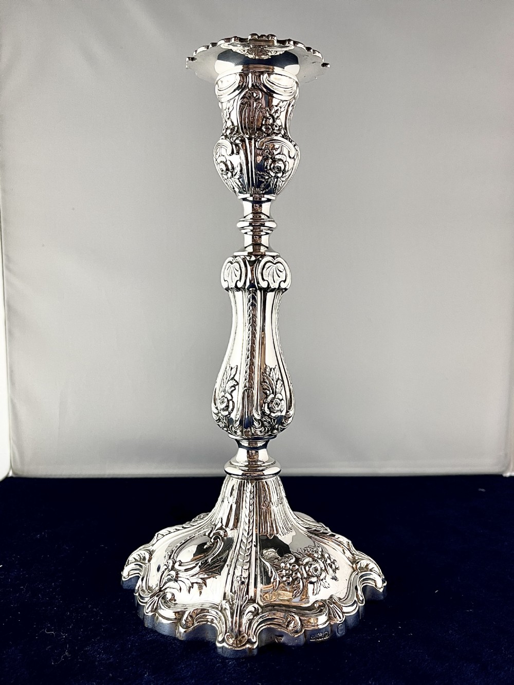 An ornate silver candlestick featuring floral repousse embossing, a contoured baluster stem, and a lobed spreading base.