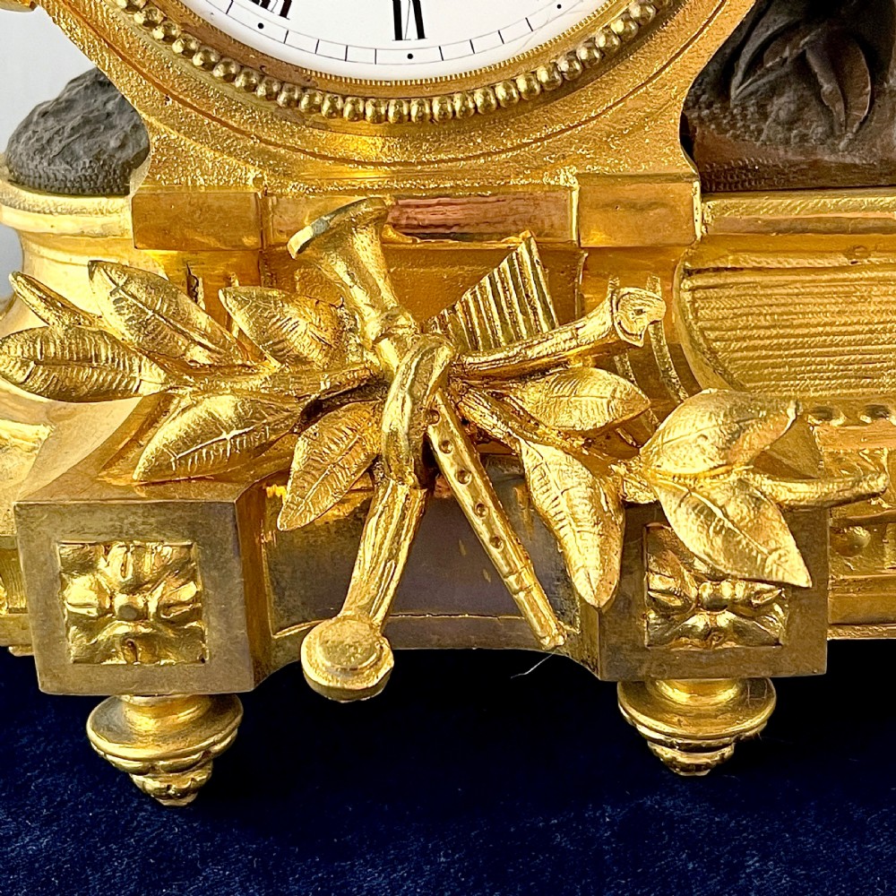 A mid-19th century French mantel clock featuring a dark patinated bronze seated figure beside a gilt bronze clock drum with a white enamel dial. View V