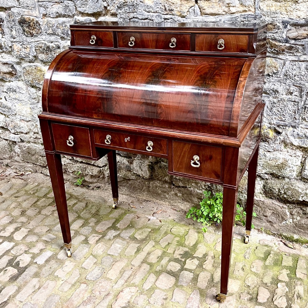 A late 19th century dark reddish-brown mahogany cylinder desk featuring a curved roll-front top, multiple drawers with brass ring handles, and tapered square legs on brass castors.