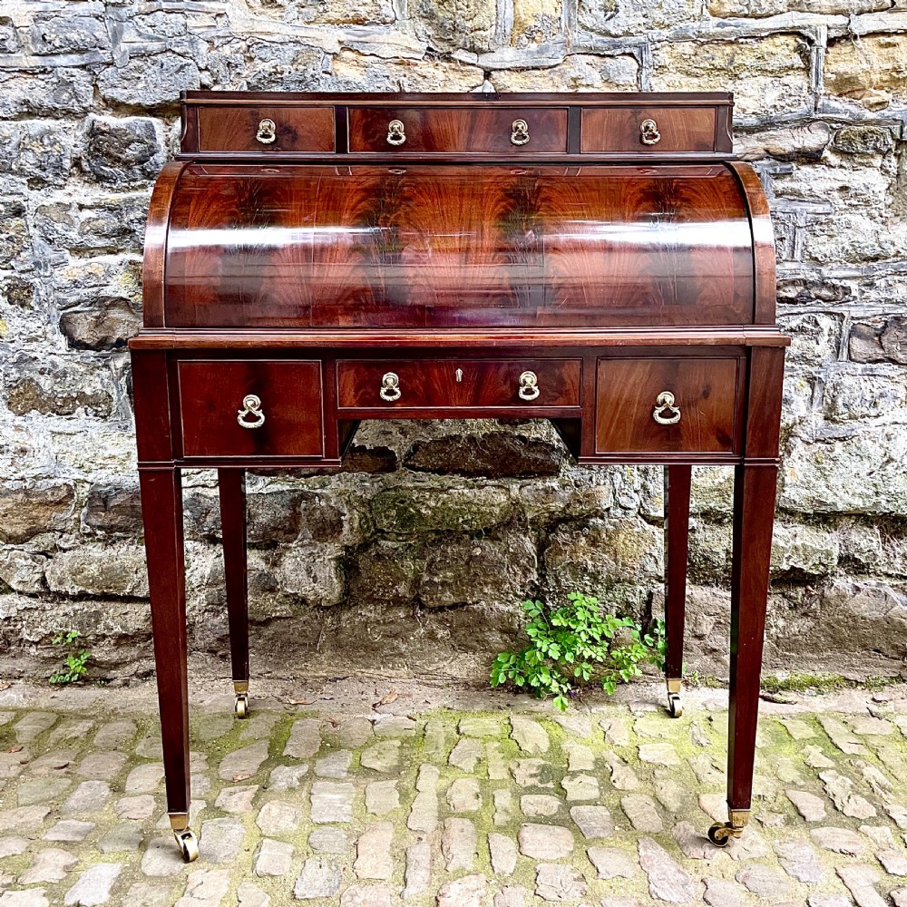 A late 19th century dark reddish-brown mahogany cylinder desk featuring a curved roll-front top, multiple drawers with brass ring handles, and tapered square legs on brass castors. View II