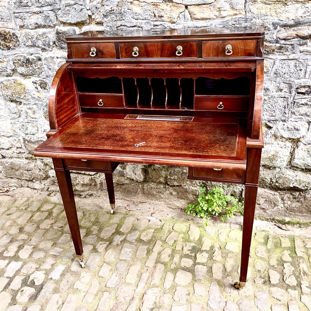 A late 19th century dark reddish-brown mahogany cylinder desk featuring a curved roll-front top, multiple drawers with brass ring handles, and tapered square legs on brass castors. View III
