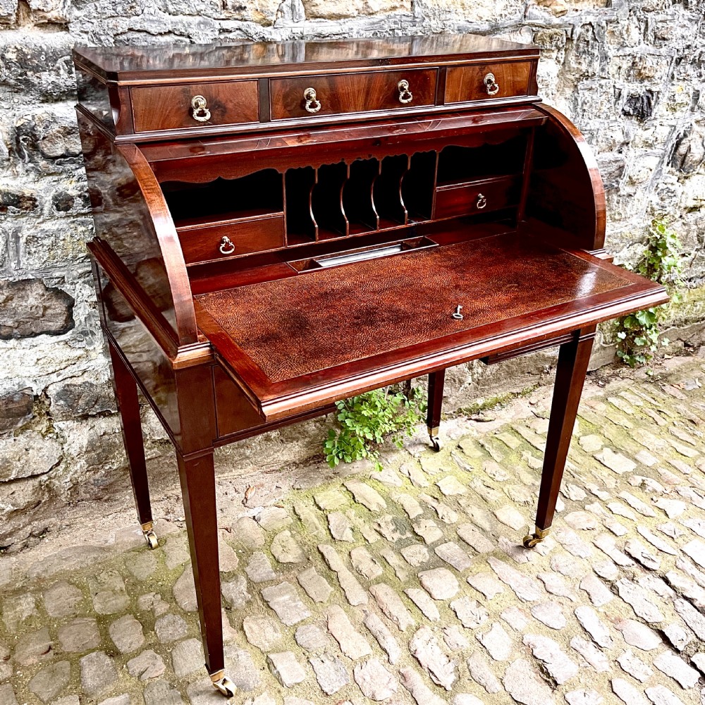 A late 19th century dark reddish-brown mahogany cylinder desk featuring a curved roll-front top, multiple drawers with brass ring handles, and tapered square legs on brass castors. View IV