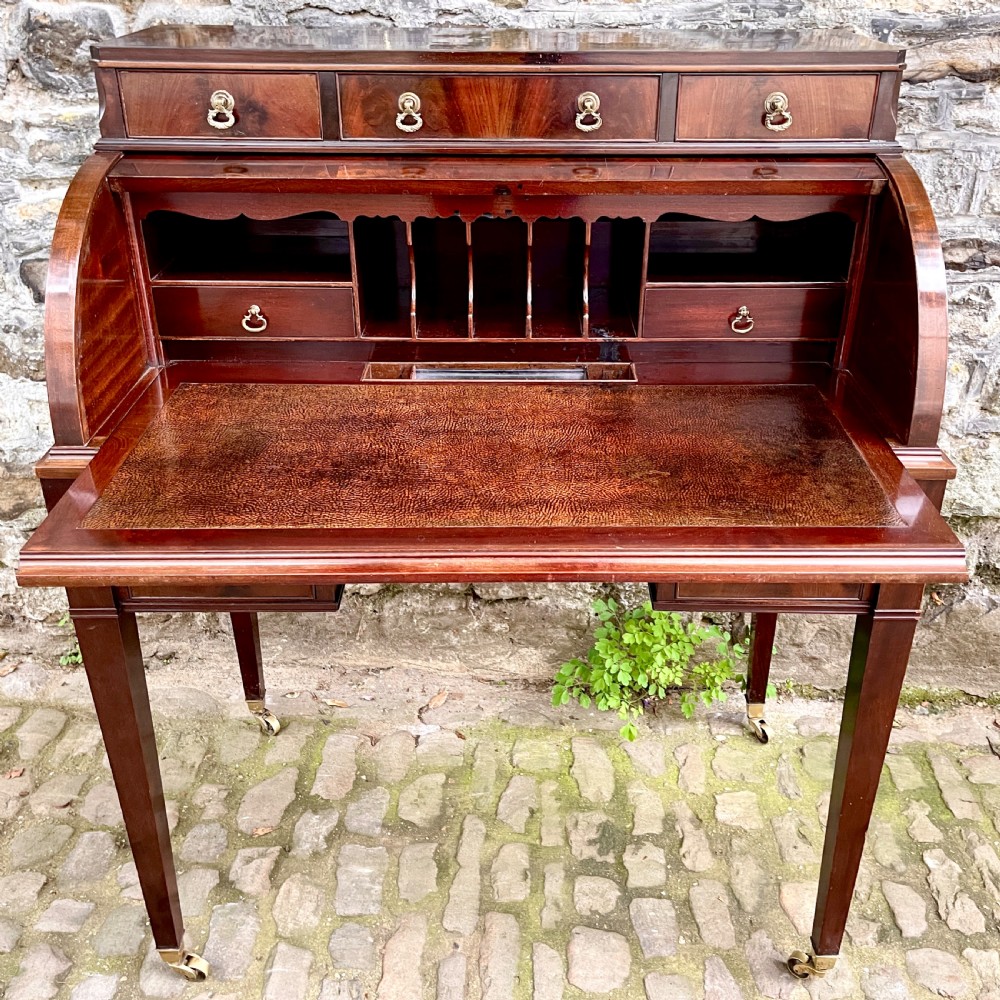 A late 19th century dark reddish-brown mahogany cylinder desk featuring a curved roll-front top, multiple drawers with brass ring handles, and tapered square legs on brass castors. View VI