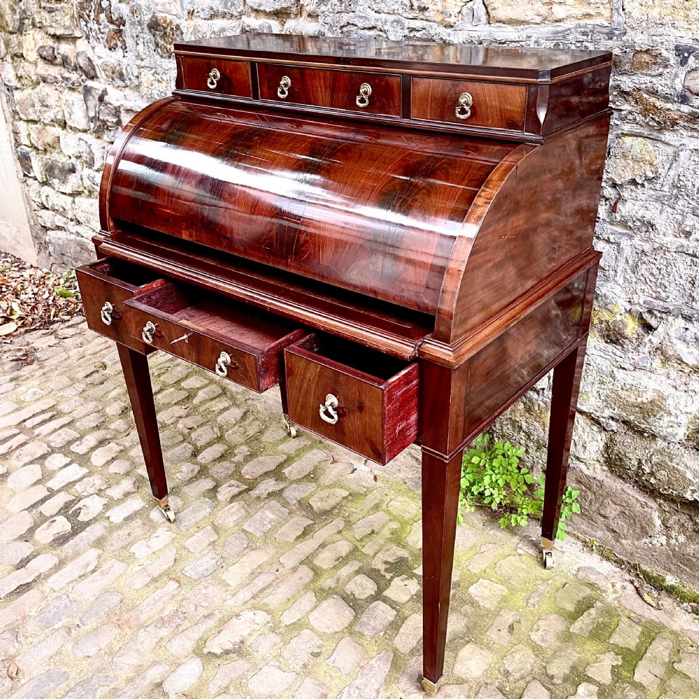 A late 19th century dark reddish-brown mahogany cylinder desk featuring a curved roll-front top, multiple drawers with brass ring handles, and tapered square legs on brass castors. View VII