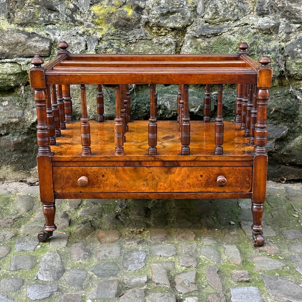 A mid-Victorian warm brown walnut Canterbury featuring turned spindles, horizontal dividers, a lower drawer with twin knobs, and turned legs on brass castors.