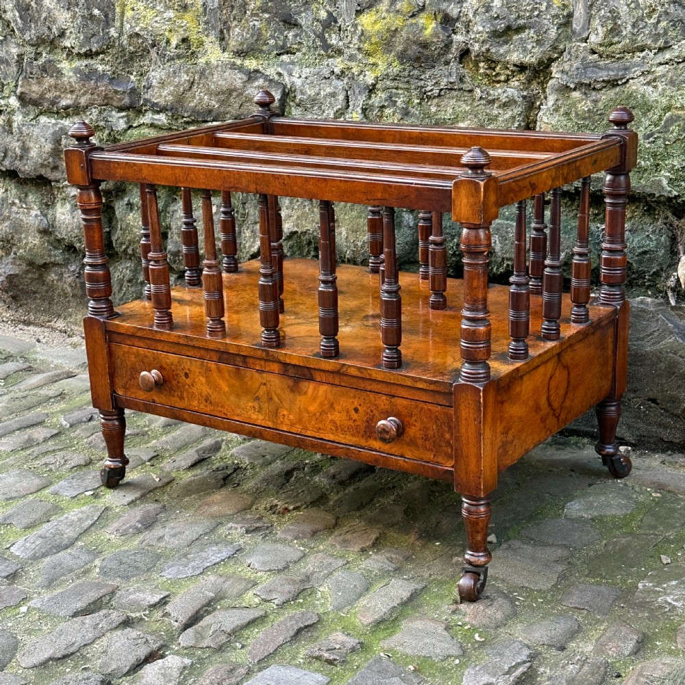 A mid-Victorian warm brown walnut Canterbury featuring turned spindles, horizontal dividers, a lower drawer with twin knobs, and turned legs on brass castors. View II