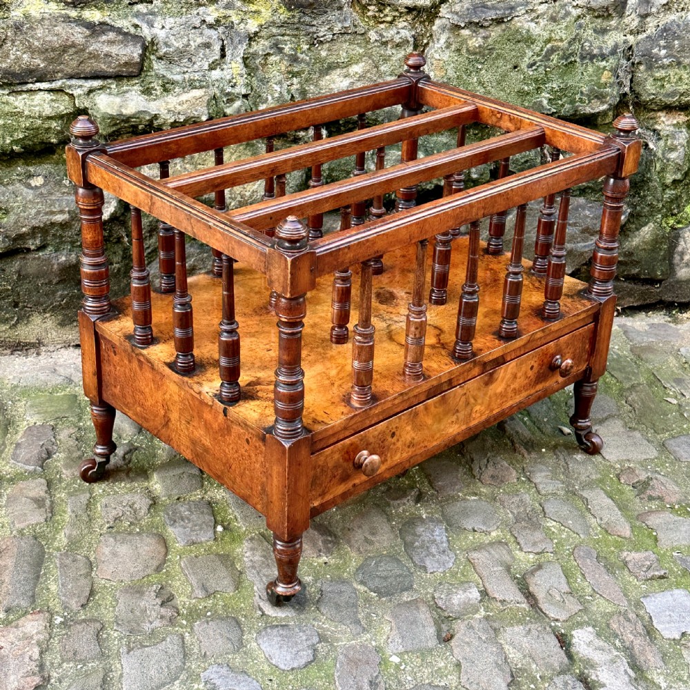 A mid-Victorian warm brown walnut Canterbury featuring turned spindles, horizontal dividers, a lower drawer with twin knobs, and turned legs on brass castors. View IV