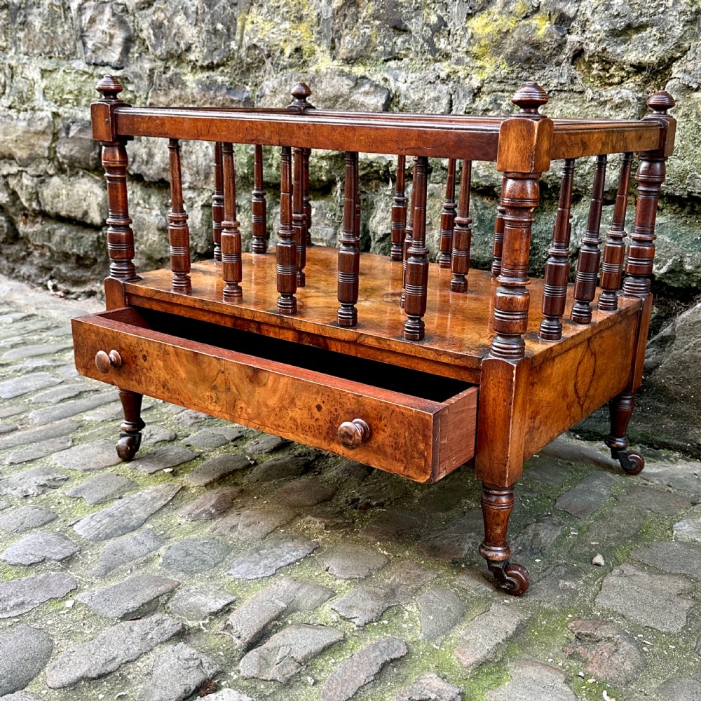 A mid-Victorian warm brown walnut Canterbury featuring turned spindles, horizontal dividers, a lower drawer with twin knobs, and turned legs on brass castors. View V