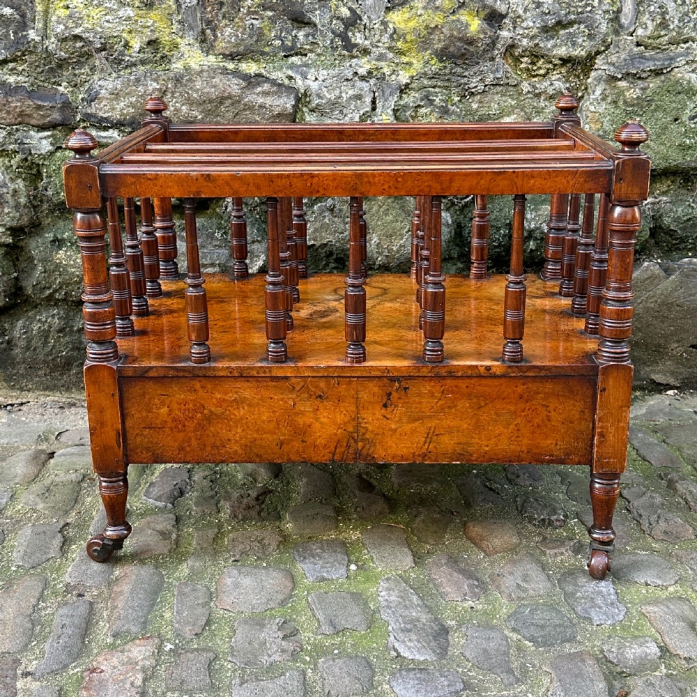 A mid-Victorian warm brown walnut Canterbury featuring turned spindles, horizontal dividers, a lower drawer with twin knobs, and turned legs on brass castors. View VII