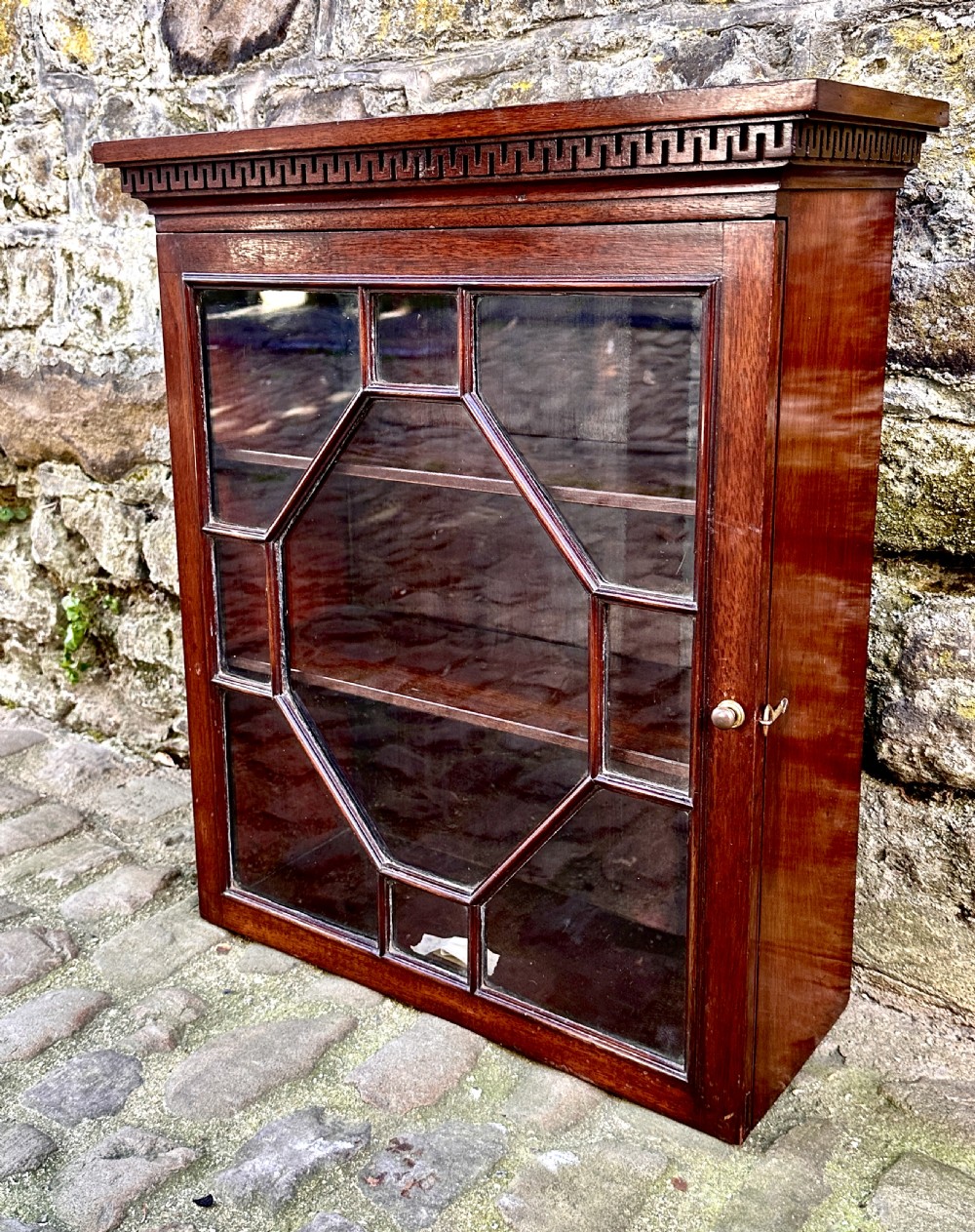 A dark mahogany Victorian wall cupboard featuring a carved Greek key frieze and a single astragal glazed door with an octagonal central glass pattern. View V