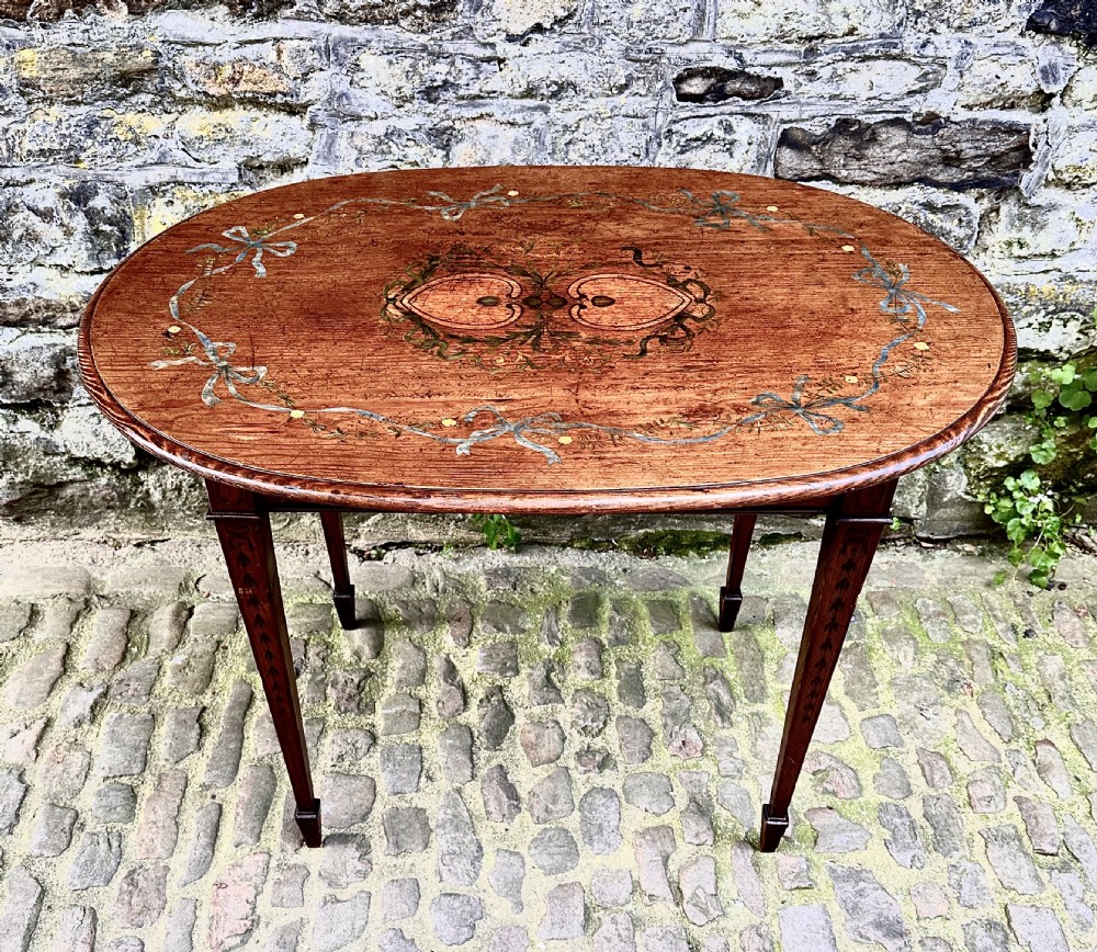 An oval wooden occasional table featuring hand-painted ribbon and floral garlands on the tabletop, supported by four tapered legs with linear details.