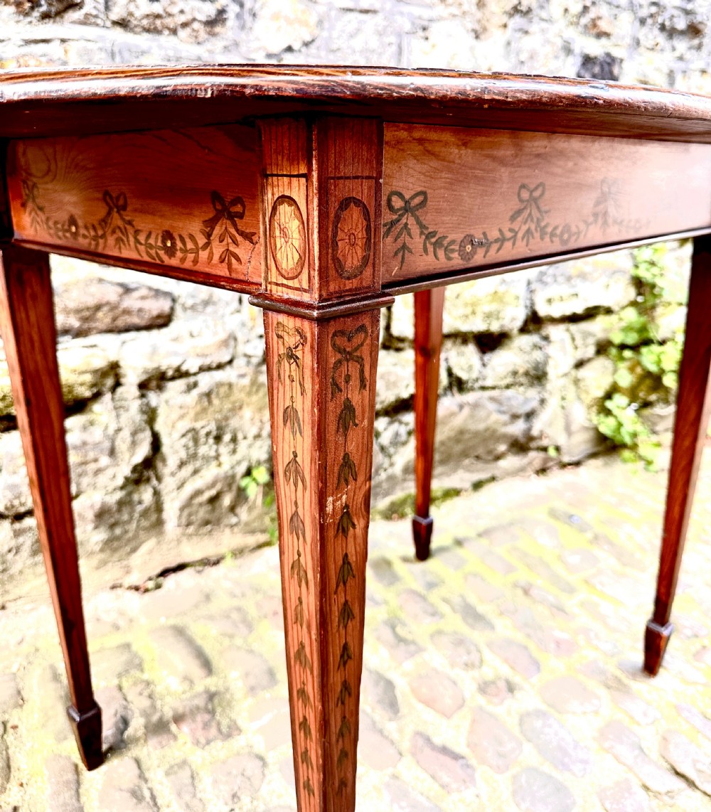 An oval wooden occasional table featuring hand-painted ribbon and floral garlands on the tabletop, supported by four tapered legs with linear details. View VI