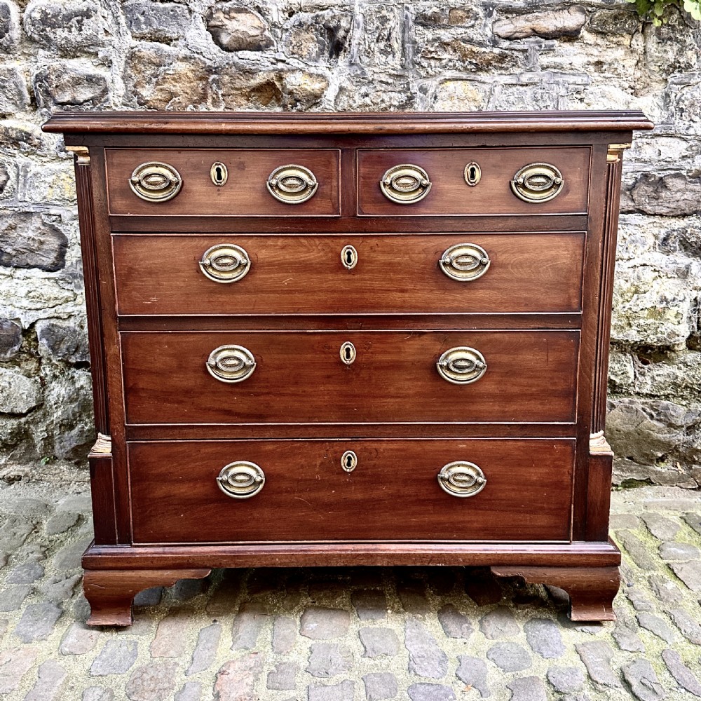 A George III mahogany chest of drawers featuring two short over three long graduated drawers, brass oval handles, fluted corner columns, and bracket feet.